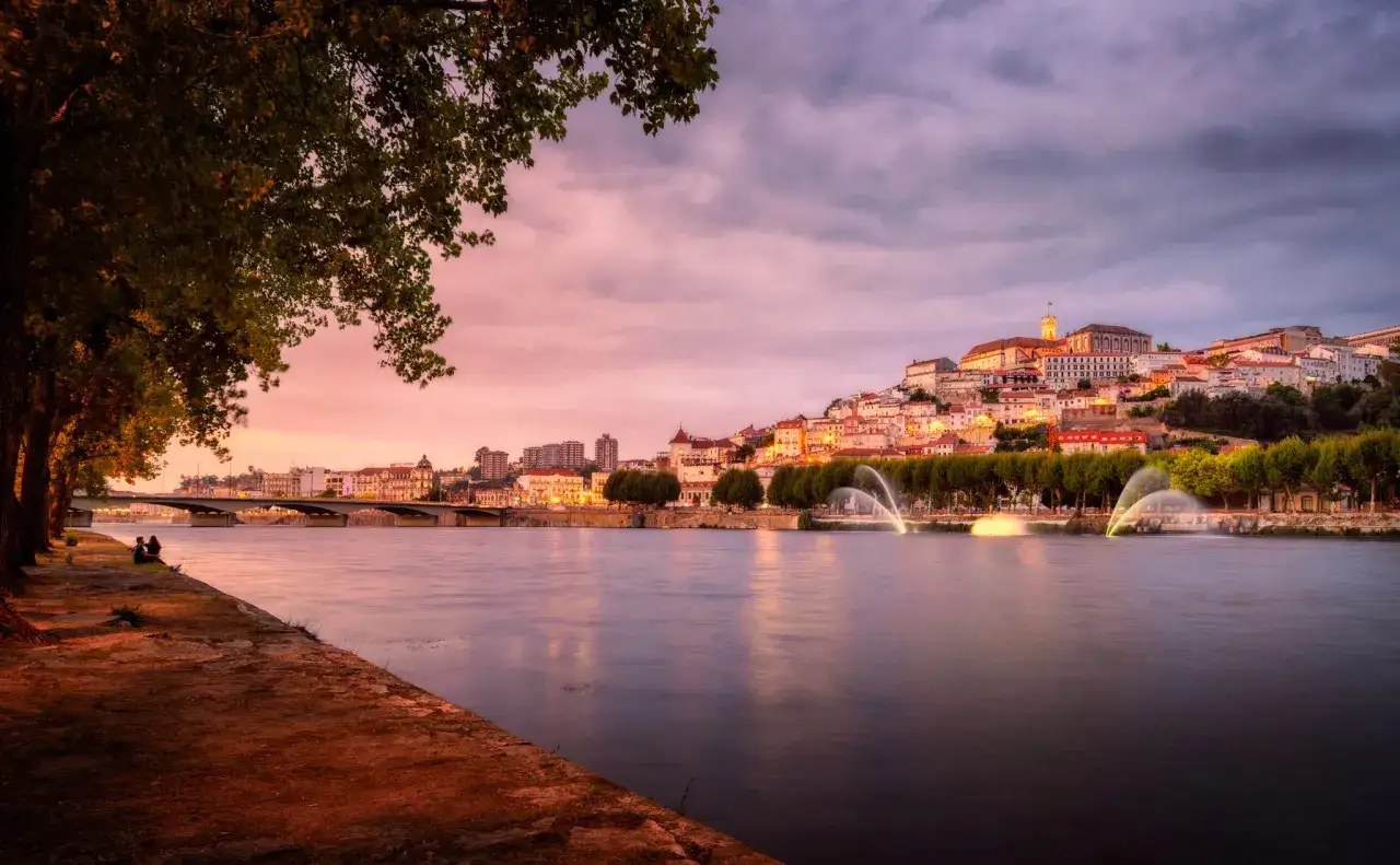 Coimbra Geheimtipps: malerische Stadtansicht am Fluss Mondego bei Sonnenuntergang mit beleuchteten Brunnen und der historischen Universität auf dem Hügel.