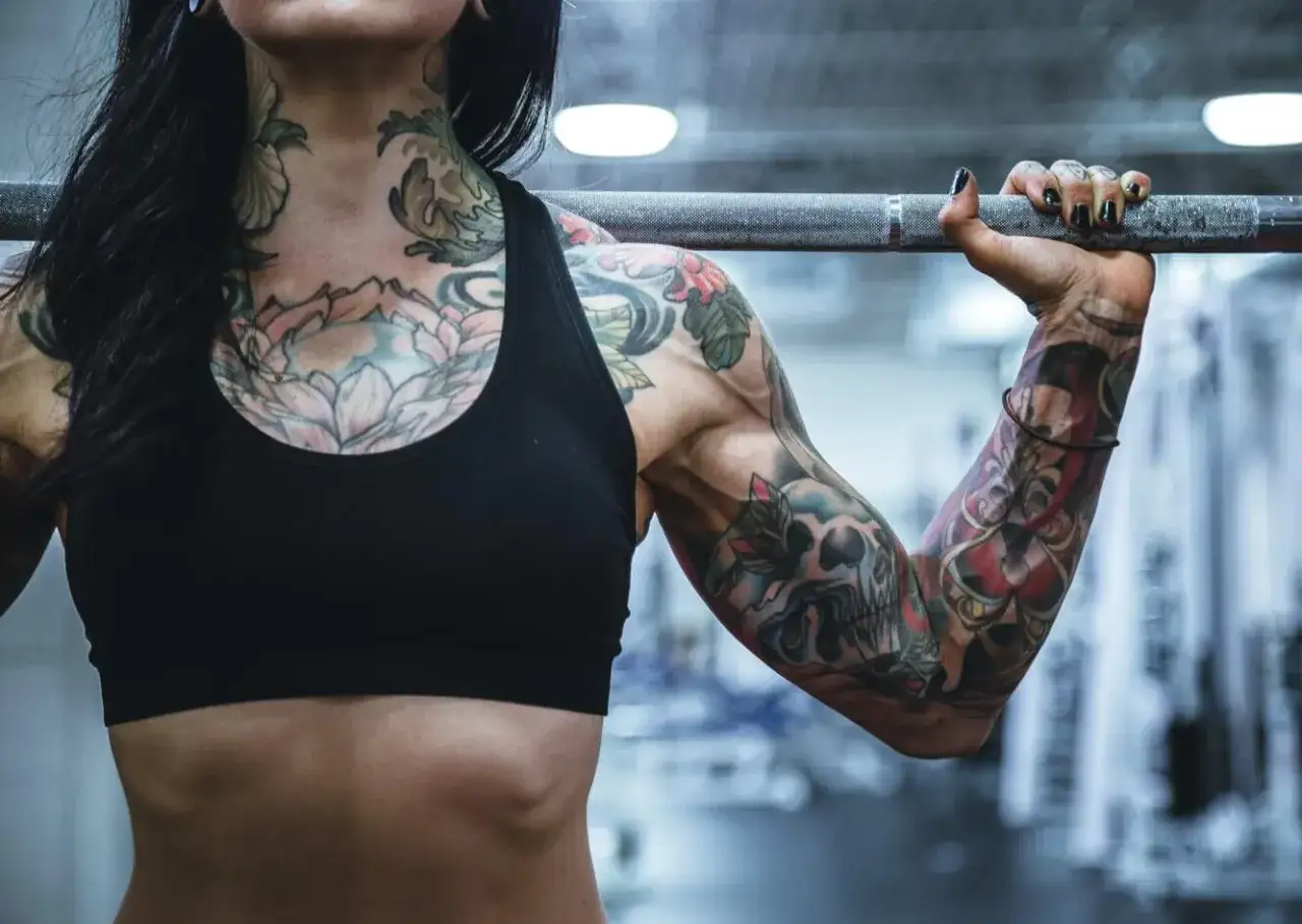 Mujer con un impresionante tatuaje en el cuello y brazos, levantando una barra. Su cuerpo fuerte muestra la dedicación al deporte y al arte corporal.