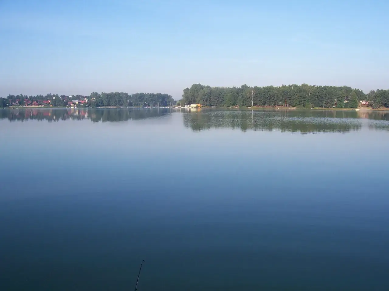 Spokojne jezioro Przeczyckie odbija błękitne niebo i zielony las. Na horyzoncie widać zabudowania.