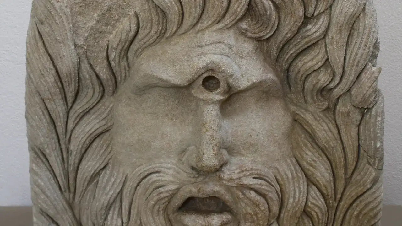 Masque de pierre d'un cyclope, avec une chevelure ondulée et une barbe fournie. Son unique œil est creusé au centre du front.
