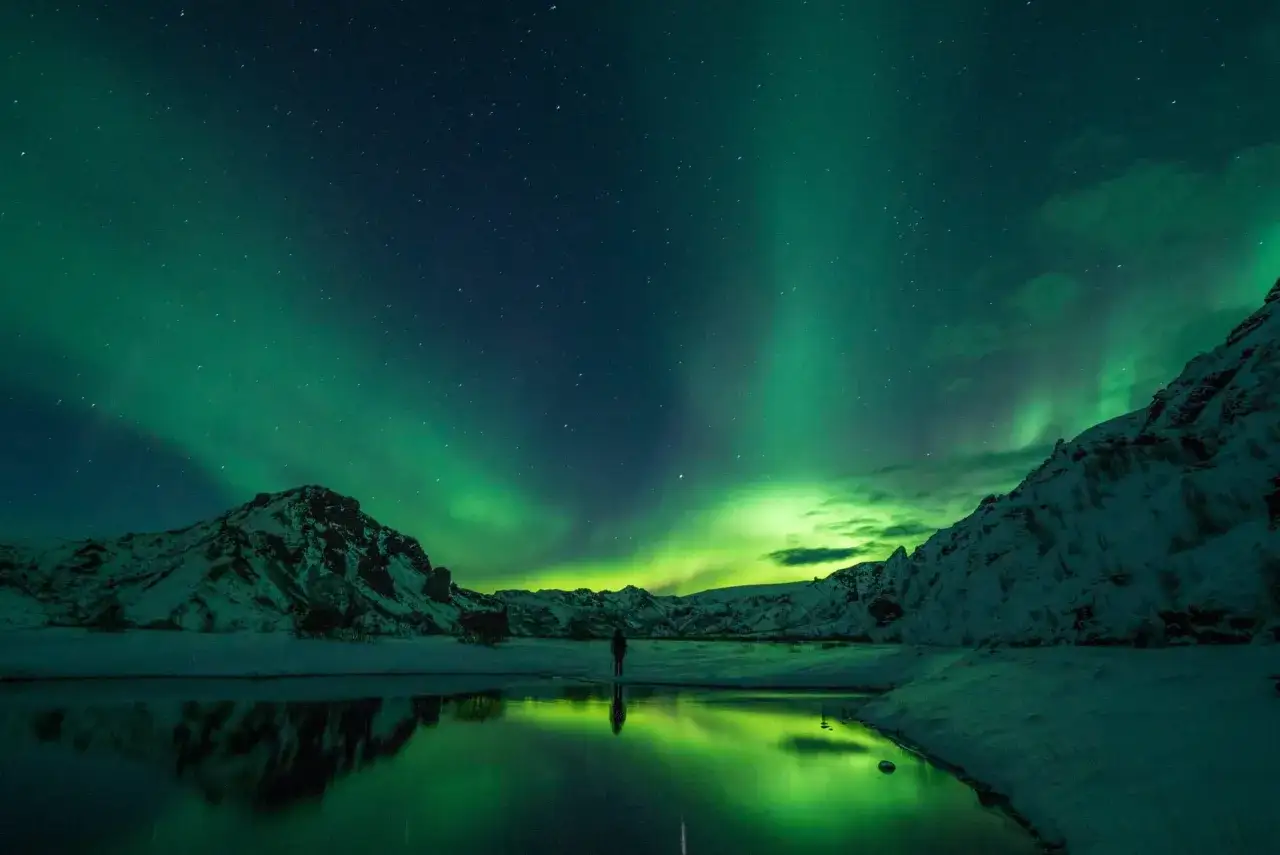 Ein einsamer Wanderer steht am Ufer eines spiegelglatten Sees und bestaunt die Nordlichter. Das Wetter auf Island im September ist perfekt f&uuml;r dieses Naturschauspiel.