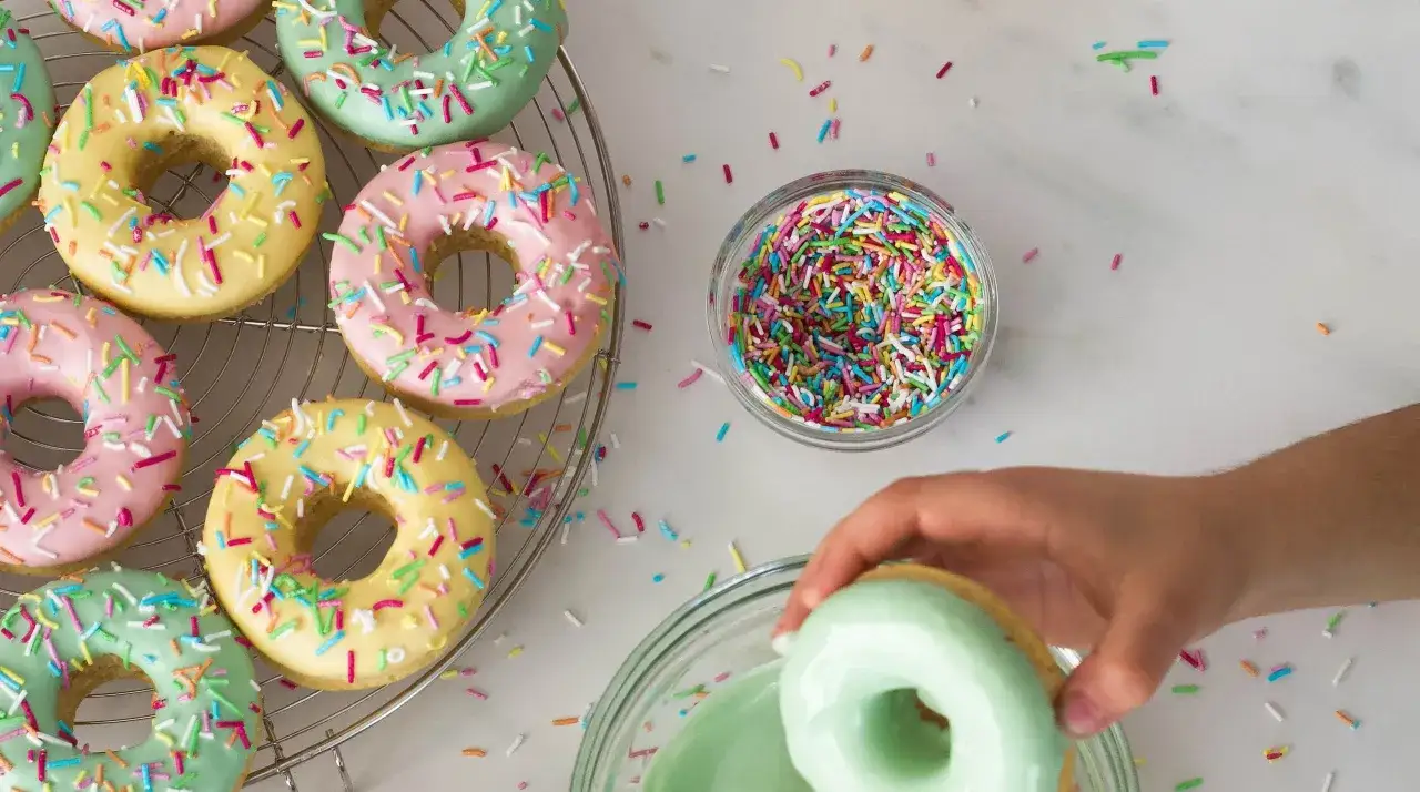 Färgglada donuts med strössel, en hand doppar en donut i grön glasyr. En härlig dag att baka donuts!