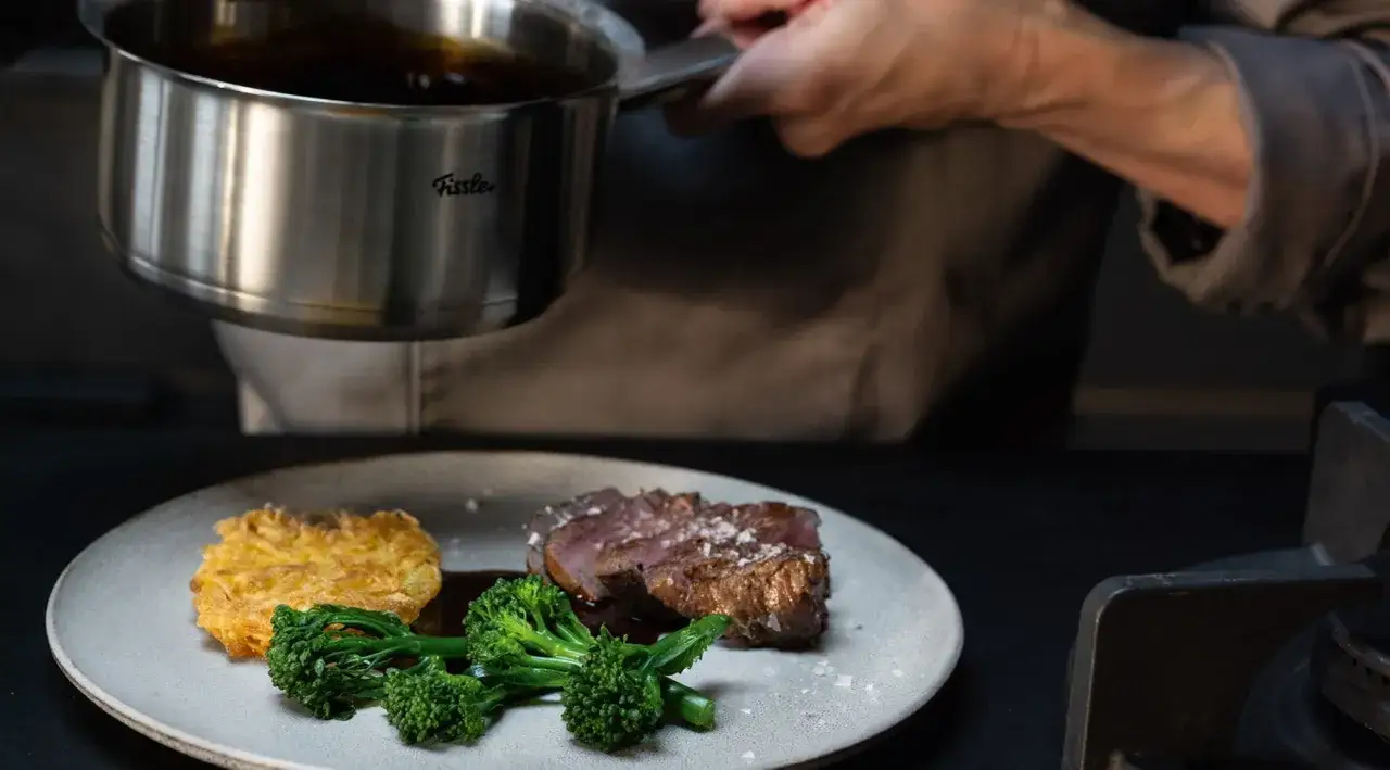 Sternekoch vollendet ein Gericht mit Portweinsauce, die aus einem Topf über ein perfekt gebratenes Steak gegossen wird. Dazu gibt es Brokkoli und Kartoffelrösti.