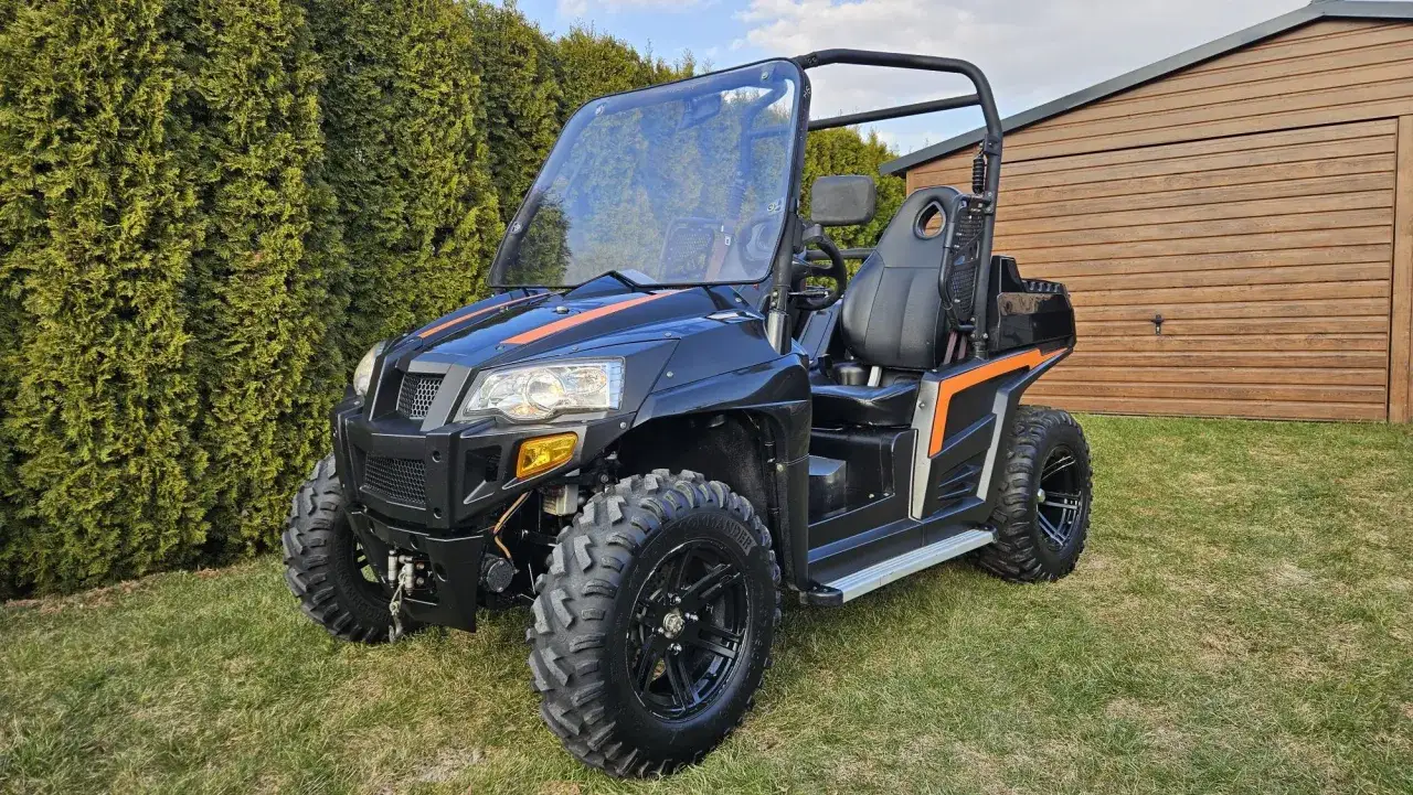 Czarny pojazd terenowy Hyosung TE 450 z pomarańczowymi akcentami stoi na trawie przed drewnianym budynkiem.