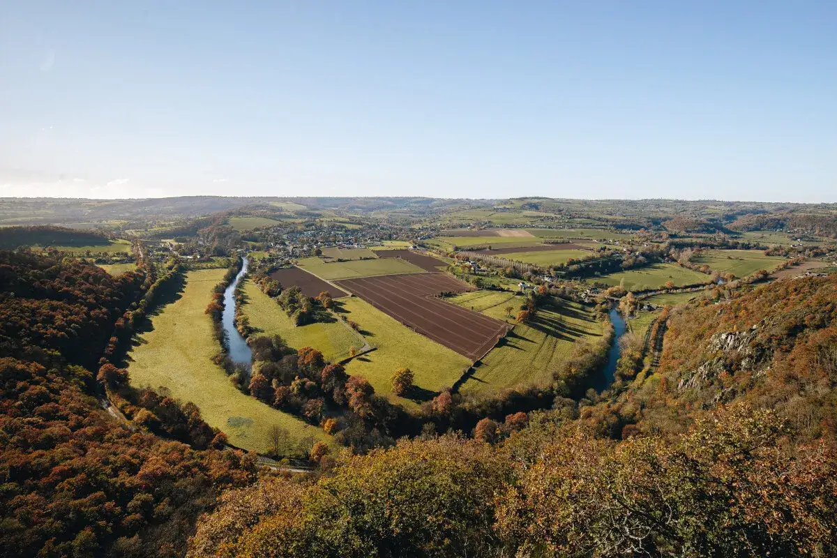 Vue panoramique de la Suisse Normande, avec une rivière sinueuse traversant des champs verdoyants et des forêts aux couleurs d'automne.