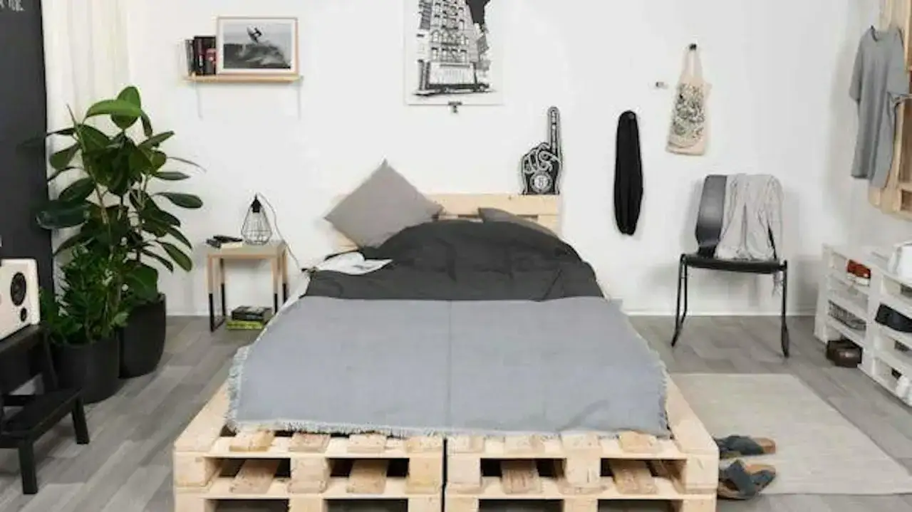 Łóżko z palet inspiracje: minimalistyczna sypialnia z drewnianą ramą łóżka, szarymi tekstyliami i rośliną doniczkową.