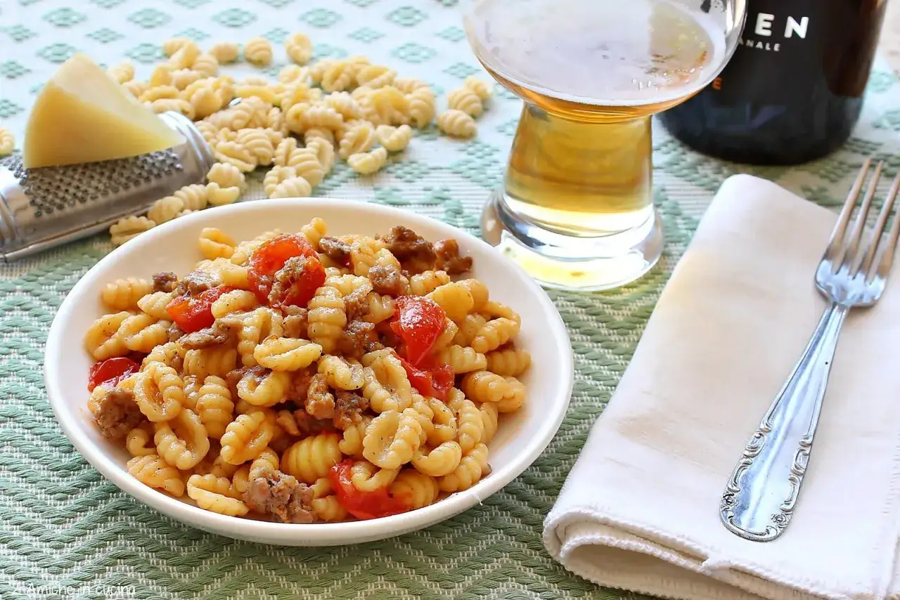 Piatto di malloreddus alla campidanese con salsiccia e pomodorini, accanto a un bicchiere di birra e formaggio grattugiato.