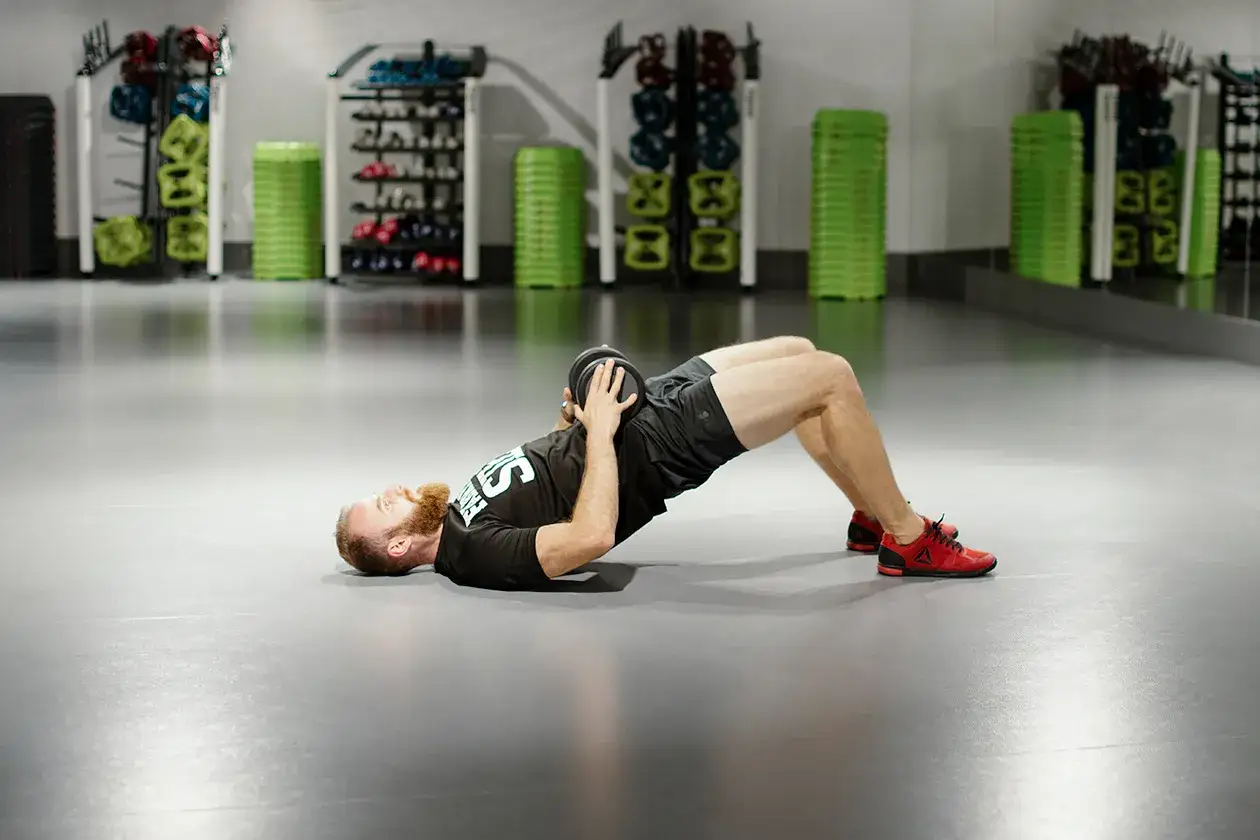 Mężczyzna z brodą wykonuje ćwiczenie glute bridge z obciążeniem na biodrach.