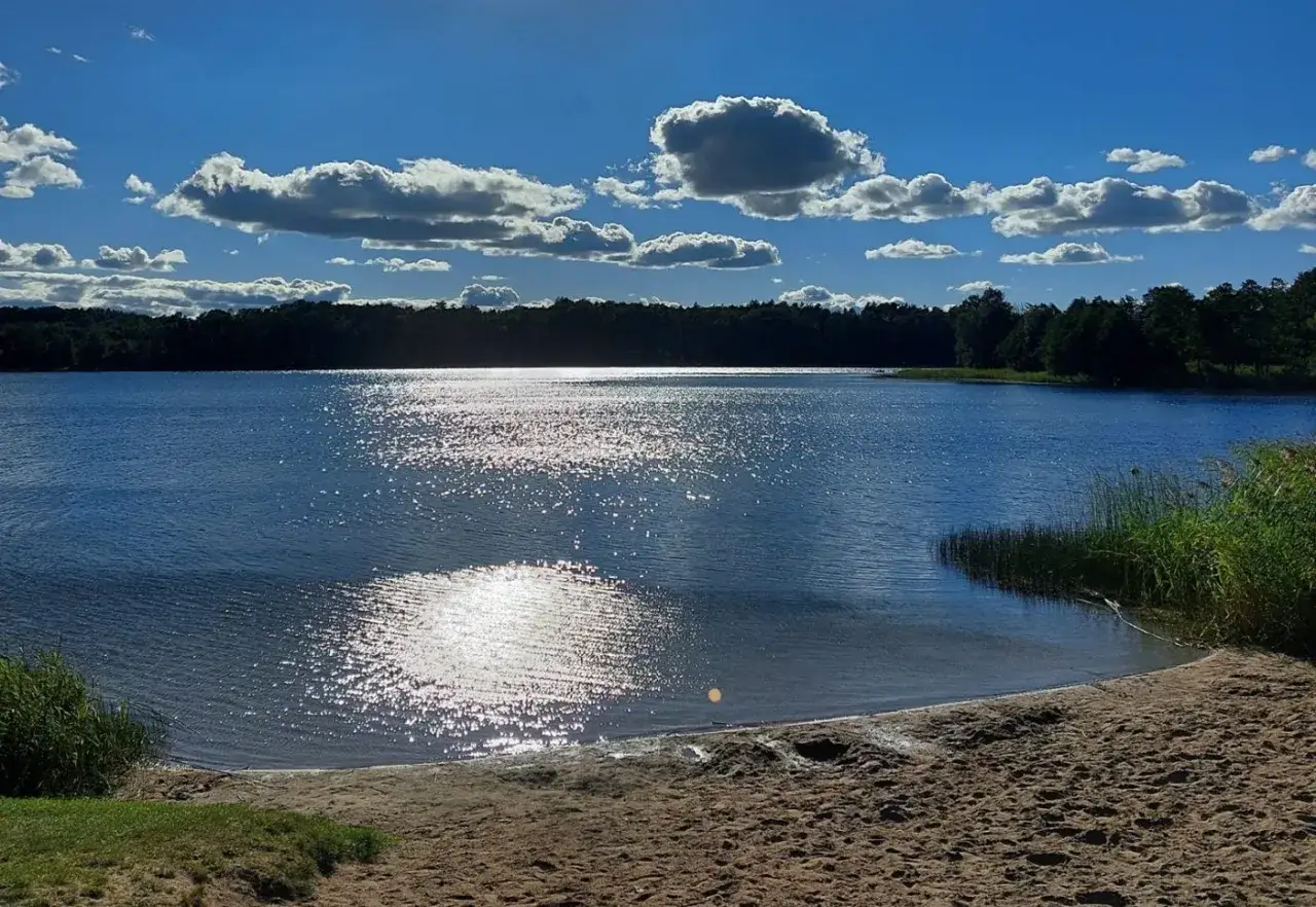 Słoneczny dzień nad jeziorem Wersminia. Błękitne niebo z białymi chmurami, lśniąca tafla wody i piaszczysta plaża zapraszają do odpoczynku.
