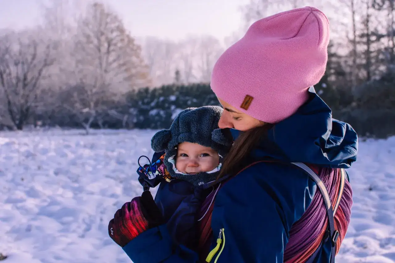 Mama w różowej czapce i granatowej kurtce nosi dziecko w nosidle. Zimowy spacer to świetna okazja, by pokazać, jak ubrać dziecko do nosidełka zimą.
