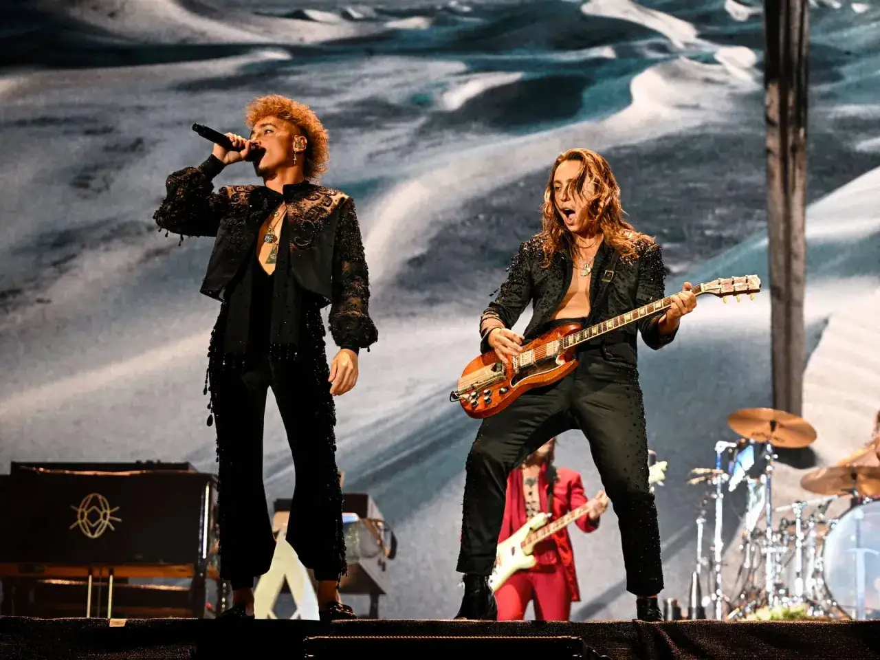 Greta Van Fleet członkowie na scenie: wokalista śpiewa do mikrofonu, gitarzysta gra na gitarze. W tle perkusja i inni muzycy.