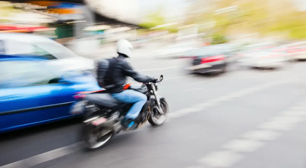 Motociclista con casco blanco adelanta por la derecha a un coche azul en una calle concurrida.