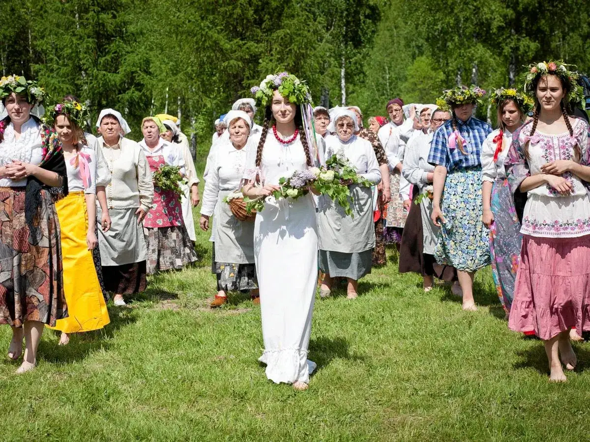 Uroczystość Zielonych Świątek. Kobiety w tradycyjnych strojach i wiankach idą przez łąkę, niosąc bukiety ziół.