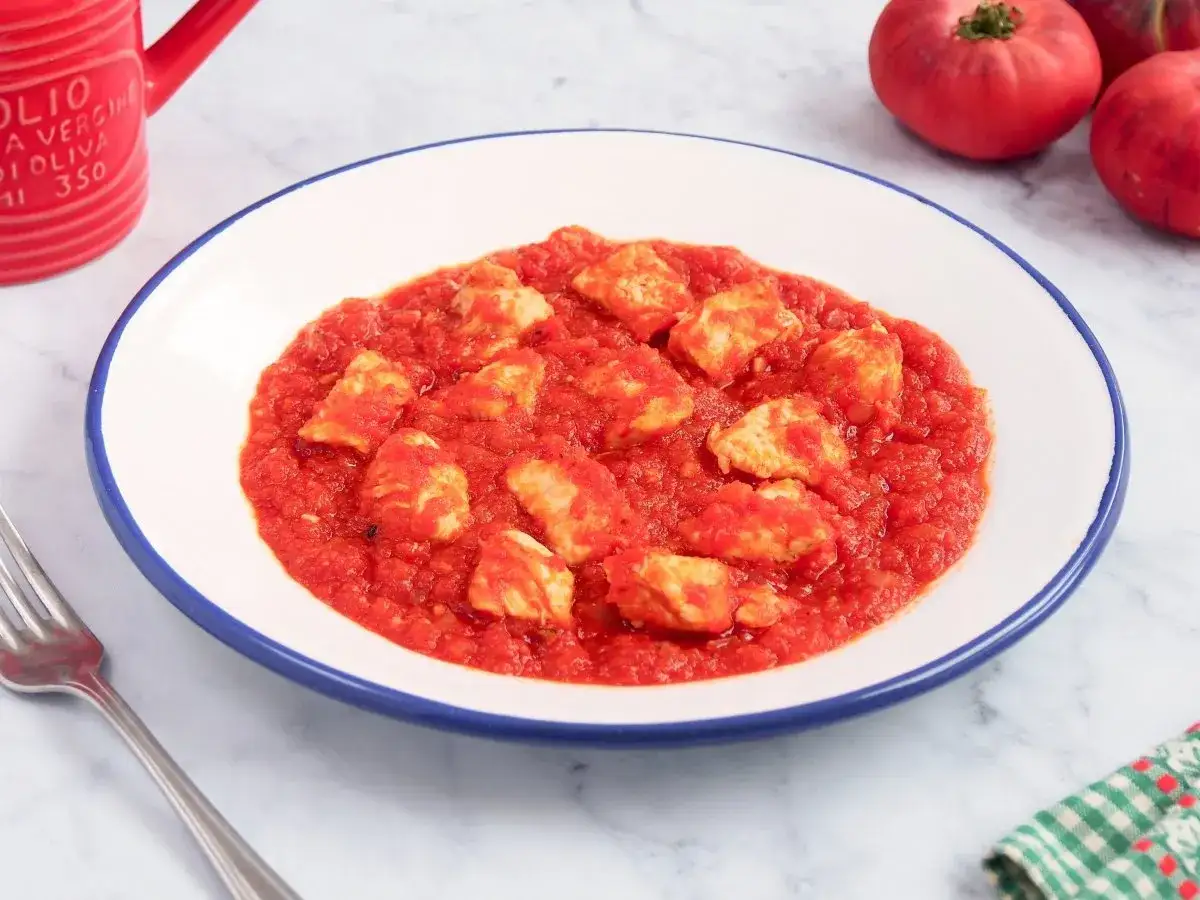 Sabroso pavo en salsa de tomate, servido en un plato blanco con borde azul. Un tenedor plateado y tomates frescos completan la escena.