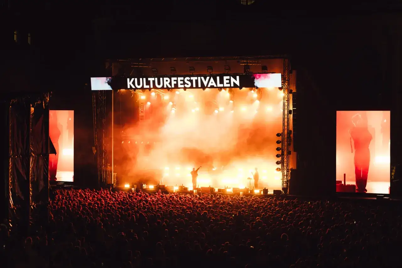 Stor publik njuter av en konsert under Kulturfestivalen. Scenen badar i orange ljus och rök, med artister som uppträder.
