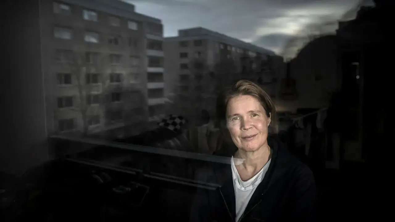Ia Genberg ler mot kameran, med en mörk bakgrund av lägenhetshus och en molnig himmel.