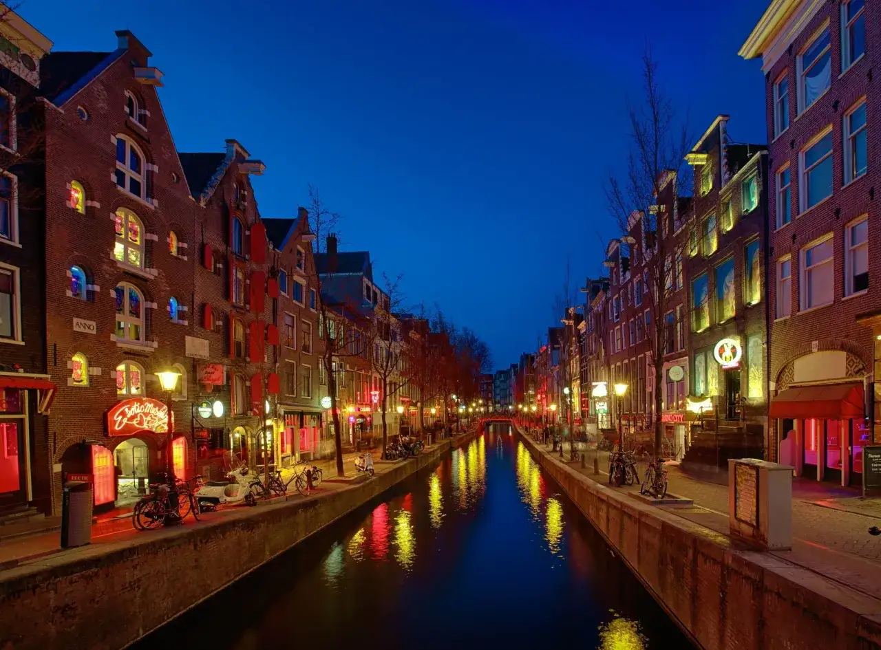 Nocny Amsterdam nad kanałem, gdzie czerwone światła latarni oświetlają fasady kamienic i odbijają się w wodzie.
