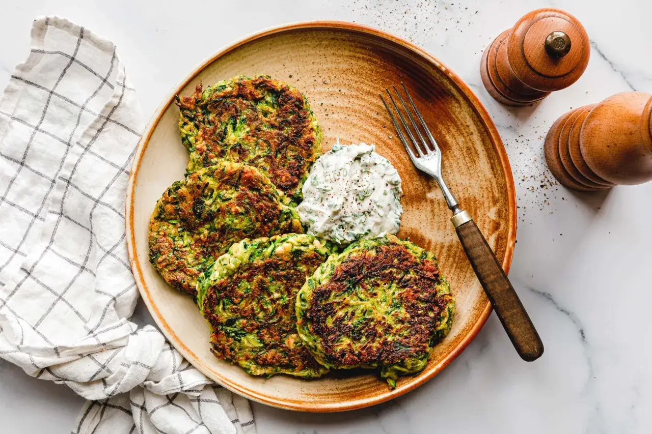 Goldbraune zucchini puffer mit einem Klecks Dip auf einem Teller, daneben eine Gabel und Gewürzstreuer.