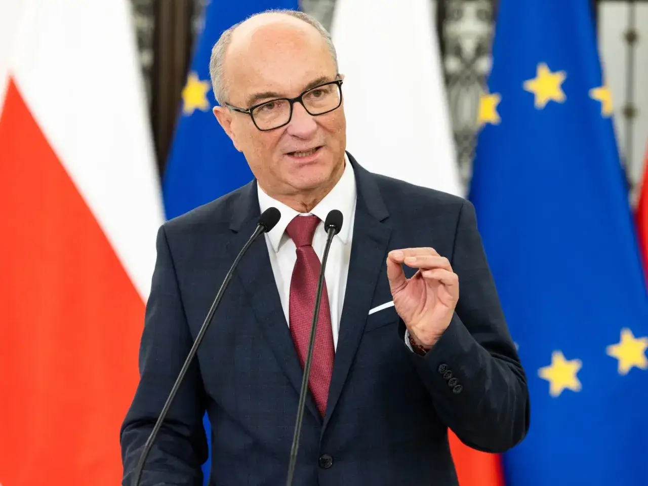 Mężczyzna w garniturze przemawia przy mikrofonach na tle flag Polski i UE. Dyskutuje o głosowaniu weta prezydenta.