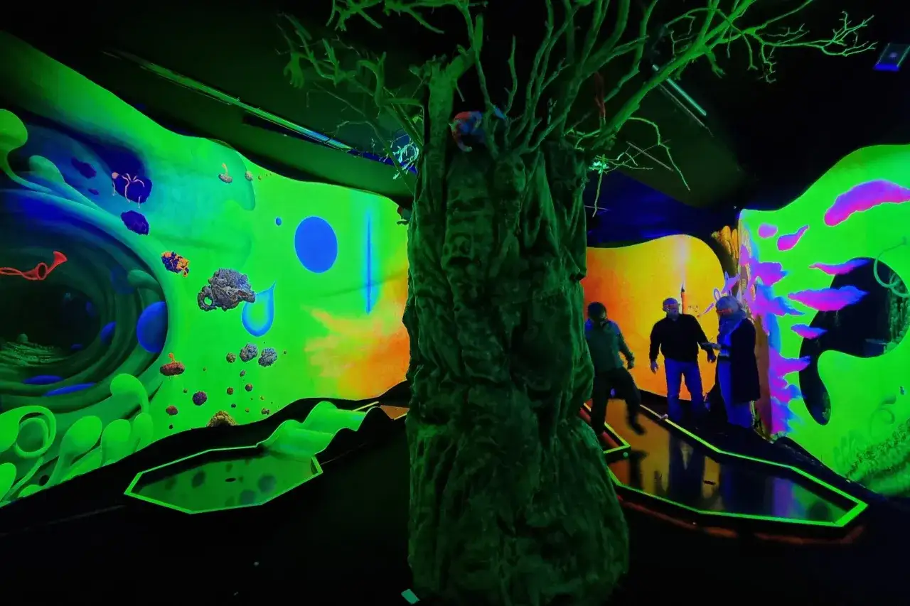 Leuchtende Farben und eine mystische Baumskulptur pr&auml;gen dieses Glow Minigolf-Erlebnis. Spieler bewegen sich durch eine neonfarbene Welt.