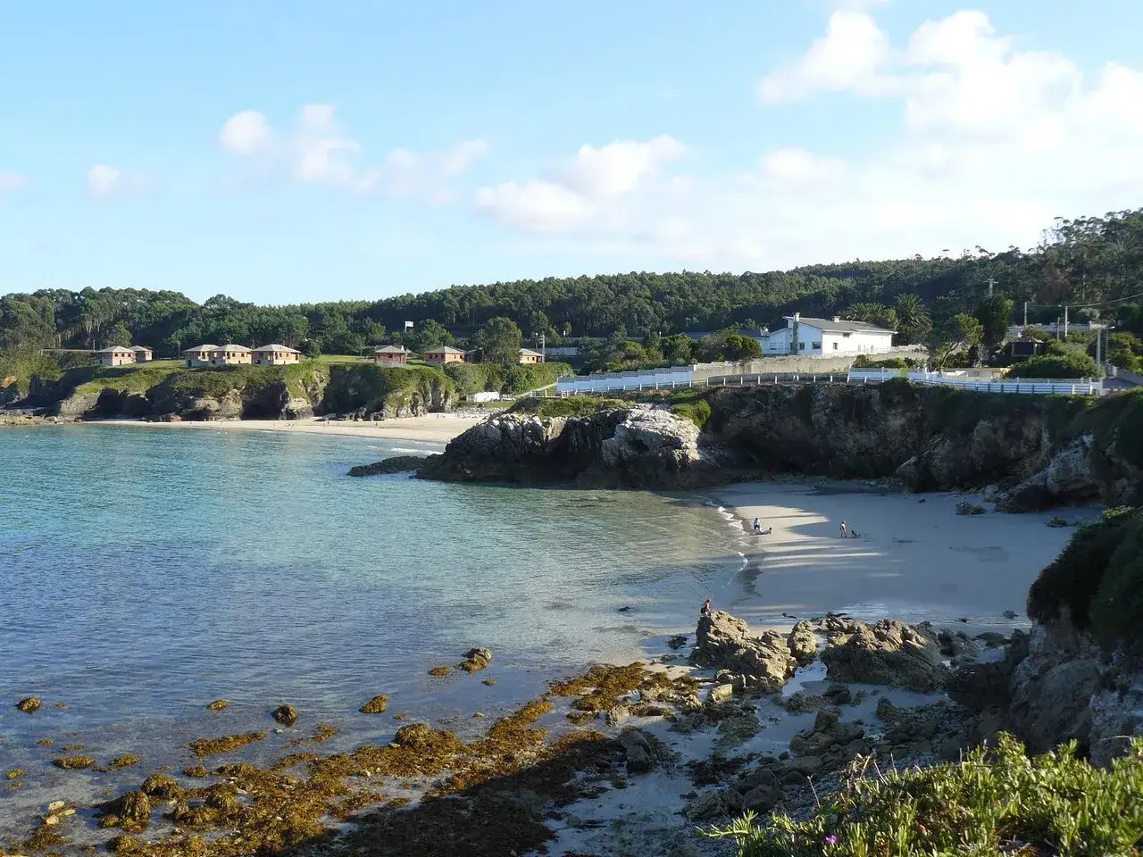 La **playa Burela** se abre a aguas cristalinas, con casitas en la colina y rocas que bordean la arena donde pasean algunas personas.