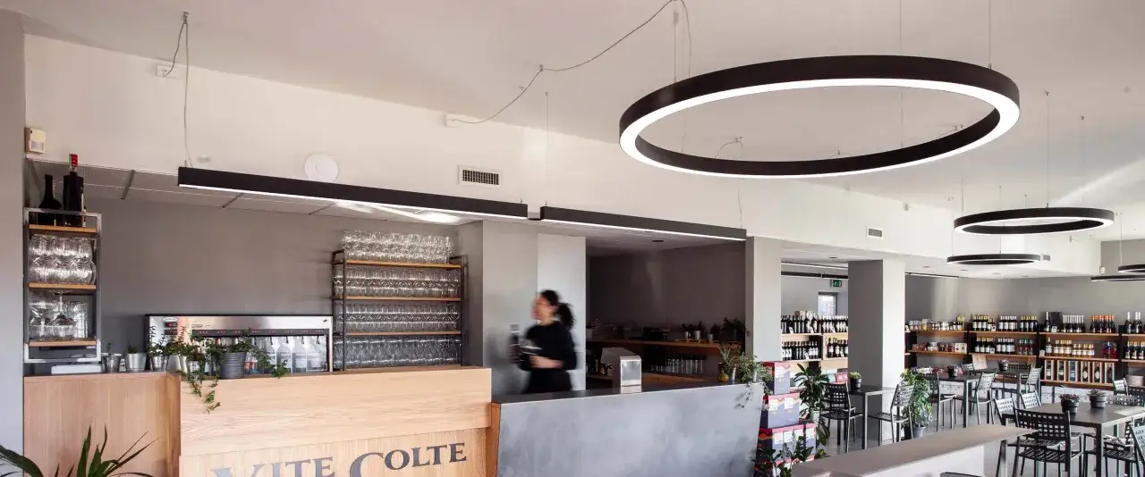 Interno di un locale moderno con bancone in legno e cemento, scaffali di bicchieri e bottiglie. Si intravede una persona che serve, in un ambiente che evoca la degustazione di **vite colte**.
