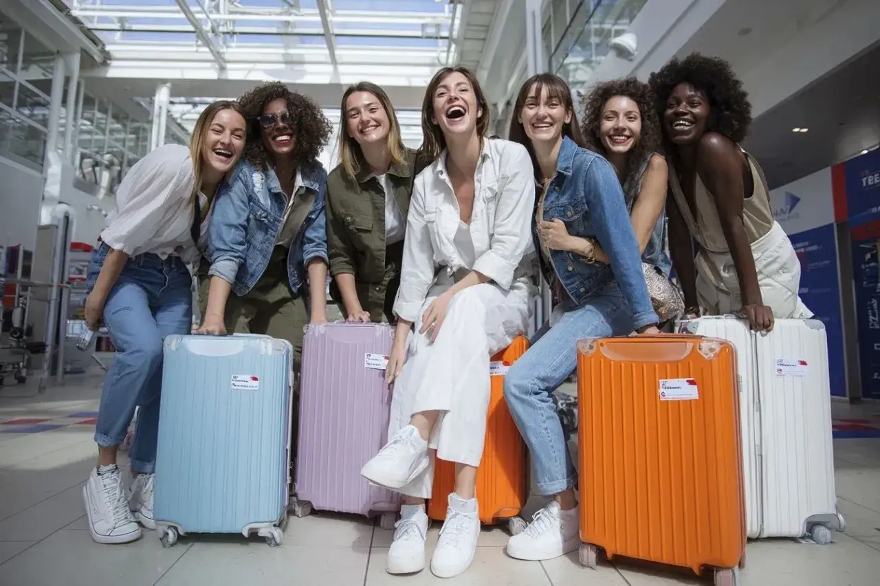 Groupe de femmes joyeuses à l'aéroport, prêtes pour leur EVJF en France. Valises colorées et sourires éclatants.