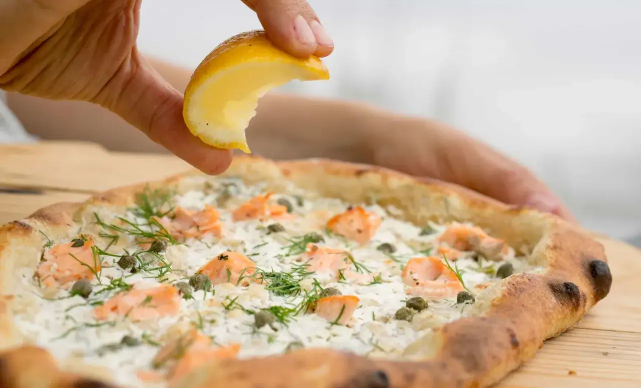 Une main presse un quartier de citron sur une pizza saumon garnie de câpres et d'aneth.