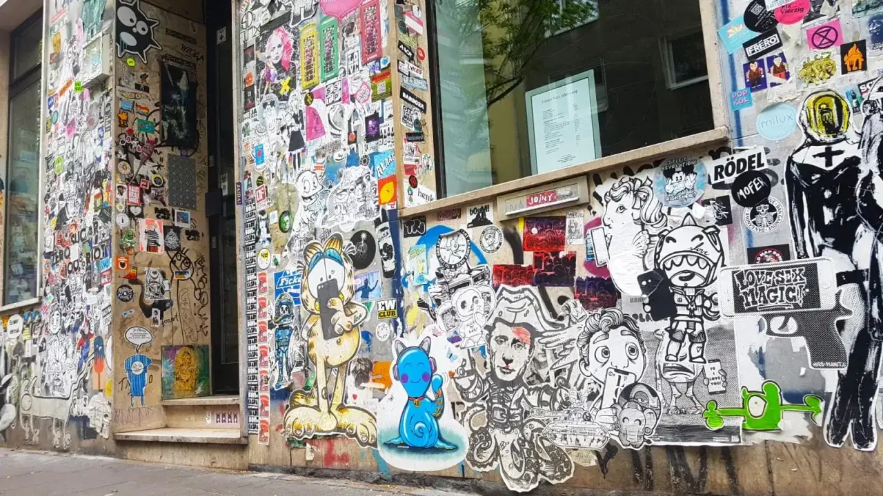 Bunte Sticker-Kunst bedeckt die Fassade eines Gebäudes im Belgischen Viertel. Cartoon-Figuren und abstrakte Designs erzählen Geschichten.