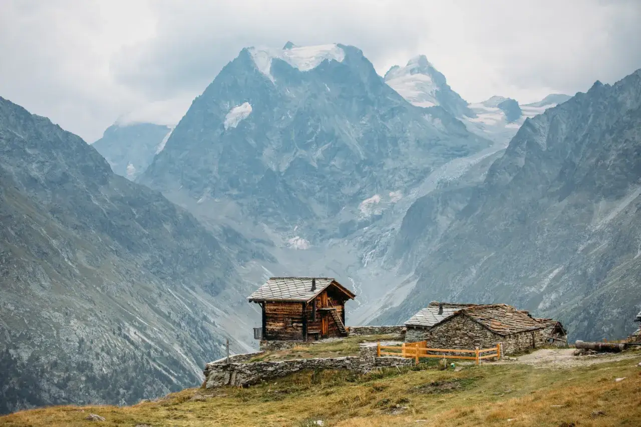 Deux chalets traditionnels en bois et pierre se dressent sur un flanc de montagne, dominant le majestueux val d'H&eacute;rens et ses sommets enneig&eacute;s.