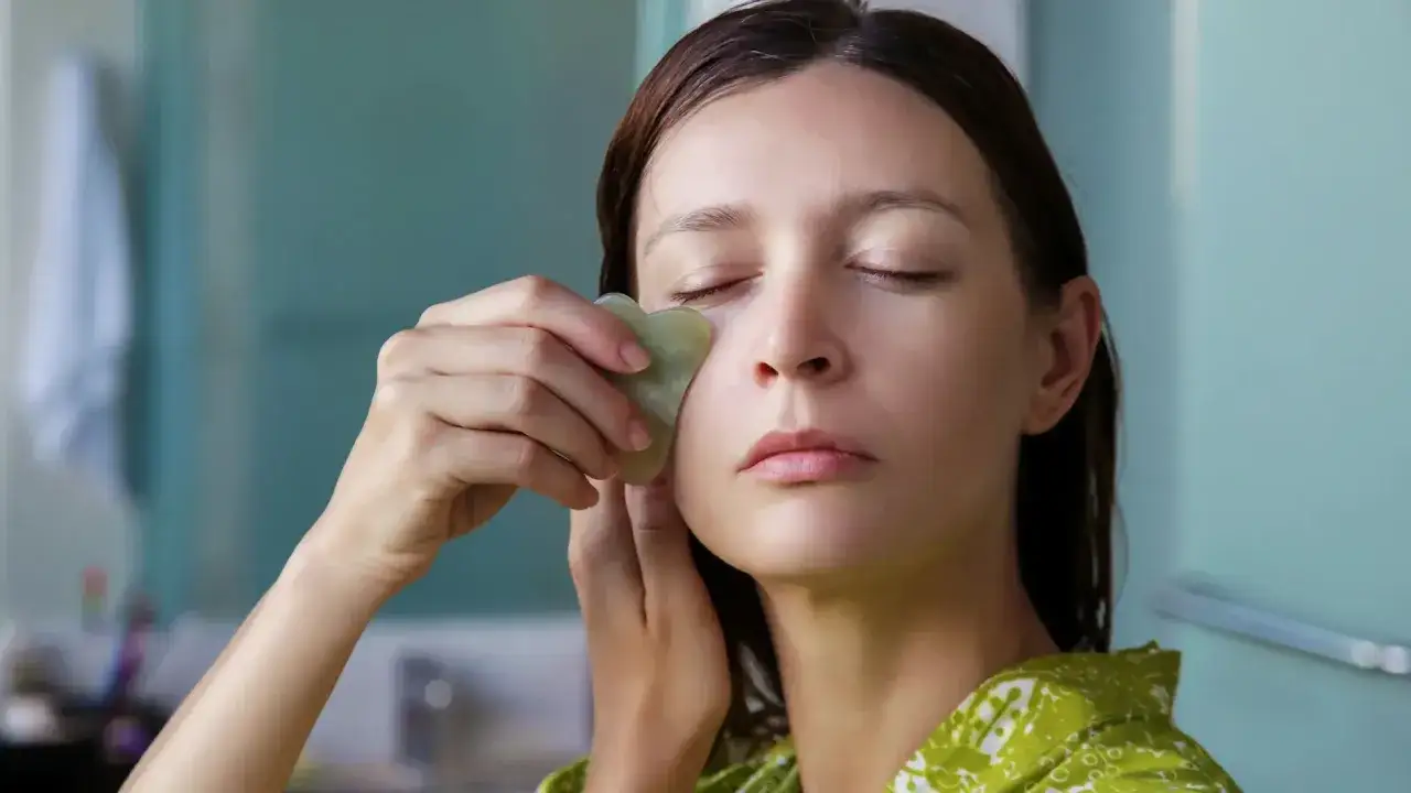 Mujer con ojos cerrados usa una piedra gua sha para masajear su rostro, aliviando la cara hinchada.