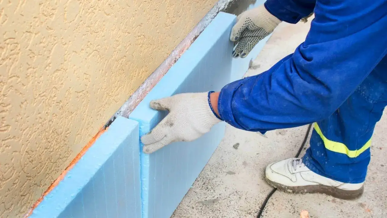 Ręce w rękawiczkach układają niebieski styropian na fundamenty przy żółtej ścianie.