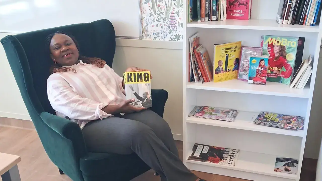 Malin Haawind sitter i en grön fåtölj och håller i en bok om Martin Luther King Jr. Bredvid henne står en bokhylla fylld med böcker och tidningar.