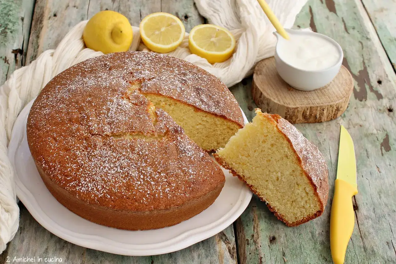 Una soffice torta allo yogurt, spolverata di zucchero a velo, con limoni freschi e una ciotolina di yogurt.