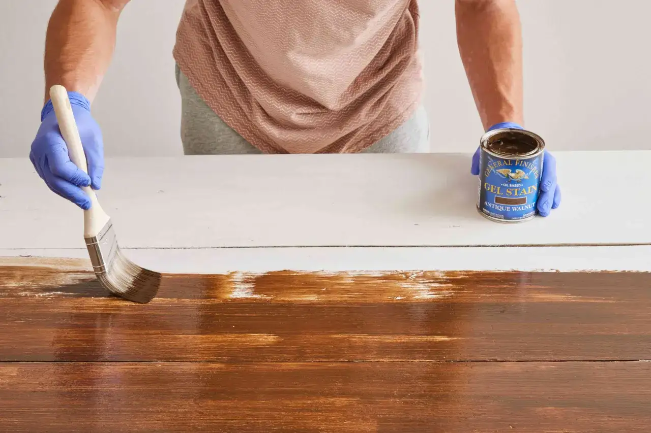 Application d'une lasure brune sur des planches de bois. Une main gantée utilise un pinceau pour étaler le produit.