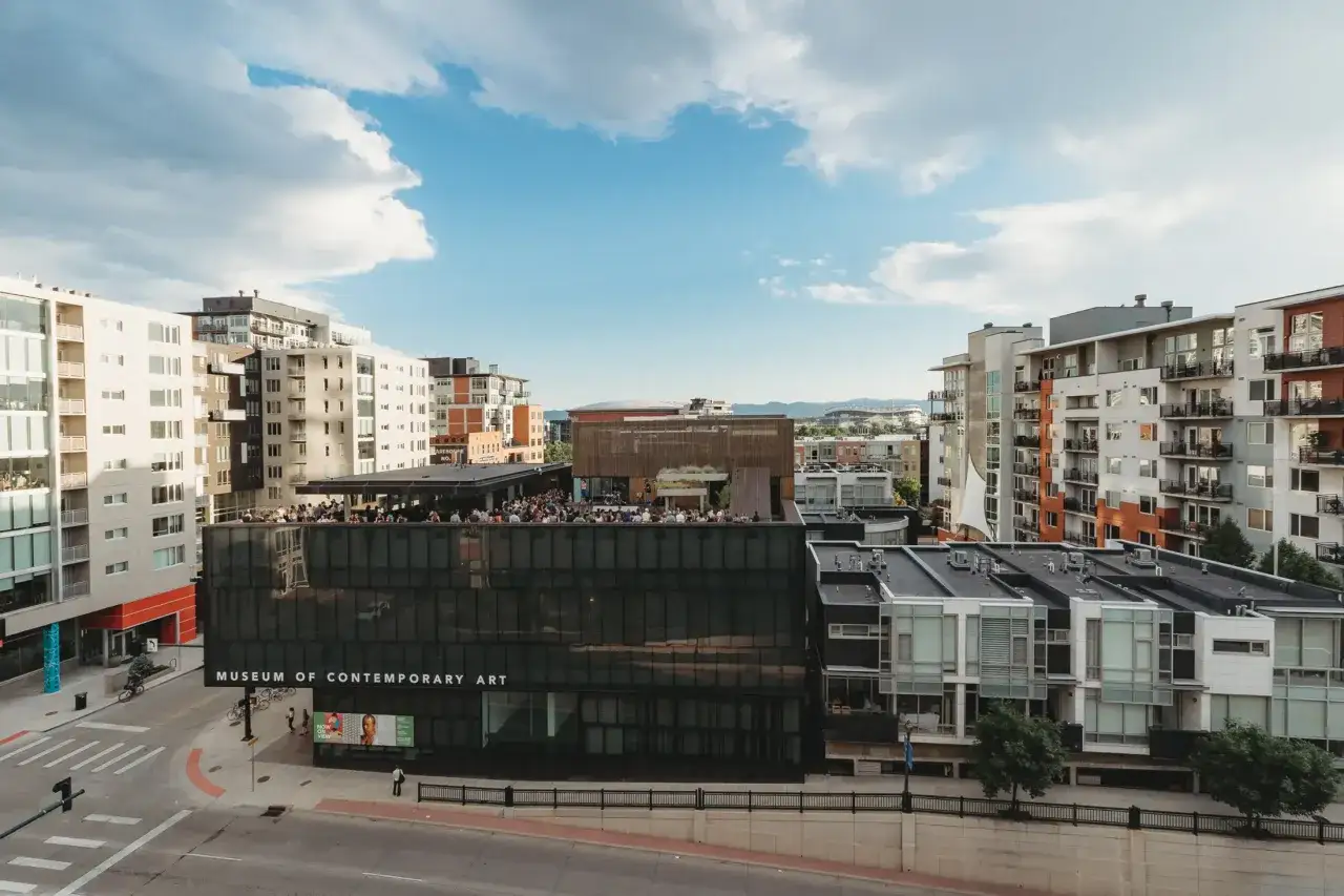 A crowd gathers on the rooftop of the Museum of Contemporary Art Denver, a modern glass building.