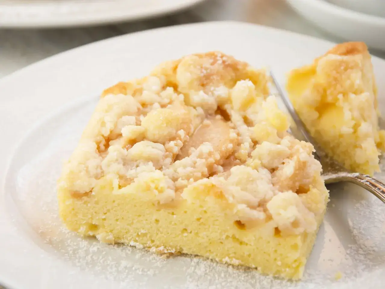 Ein St&uuml;ck von Omas Apfelkuchen mit Streuseln, best&auml;ubt mit Puderzucker, auf einem wei&szlig;en Teller.
