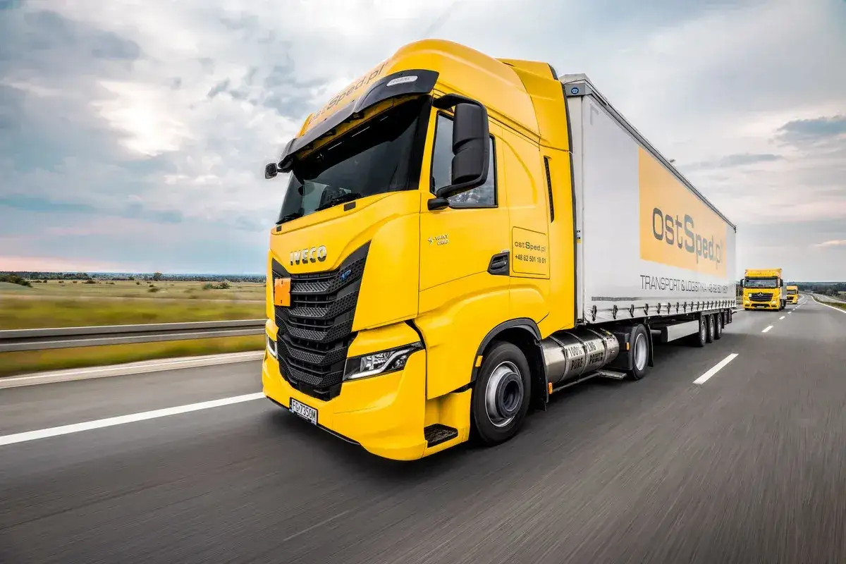 Żółta ciężarówka Iveco z naczepą OstSped.pl na autostradzie. Praca solówka międzynarodówka w ruchu.