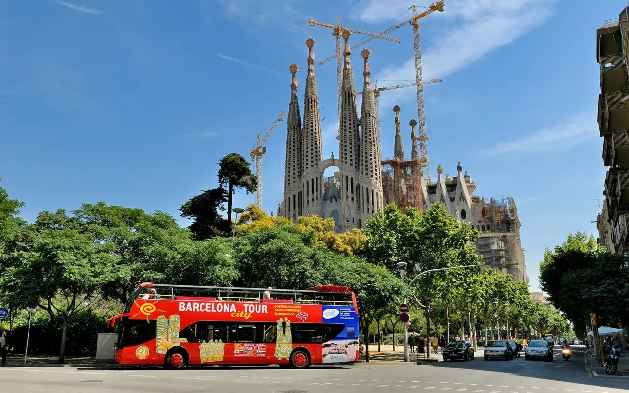 A red hop on hop off bus in Spain passes by the Sagrada Familia, a stunning basilica under construction, with cranes reaching towards the blue sky.