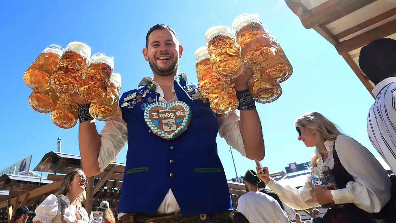 Uśmiechnięty mężczyzna w tradycyjnym stroju niesie siedem kufli piwa na Oktoberfest 2025.