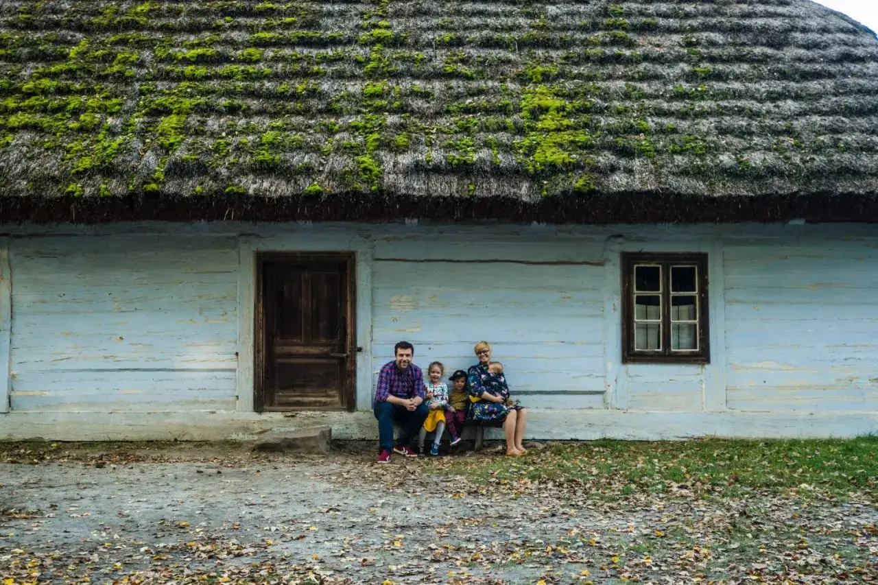 Rodzina siedzi przed drewnianym domem z zielonym, omszałym dachem. Wspaniałe atrakcje w promieniu 100 km od Radomia czekają na odkrycie!