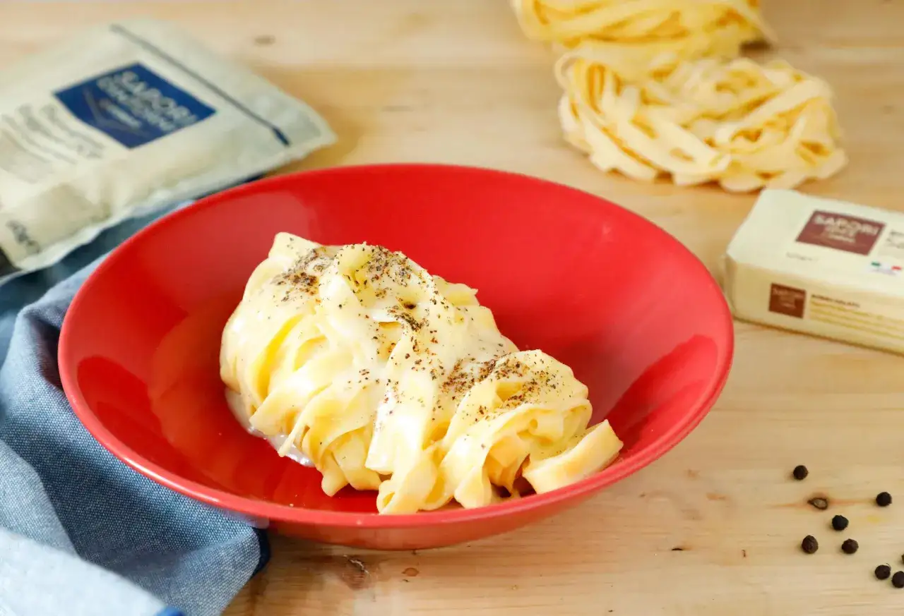 Un piatto di fettuccine alfredo fumanti, condite con una cremosa salsa al parmigiano e pepe nero. Accanto, ingredienti per preparare questo classico piatto.