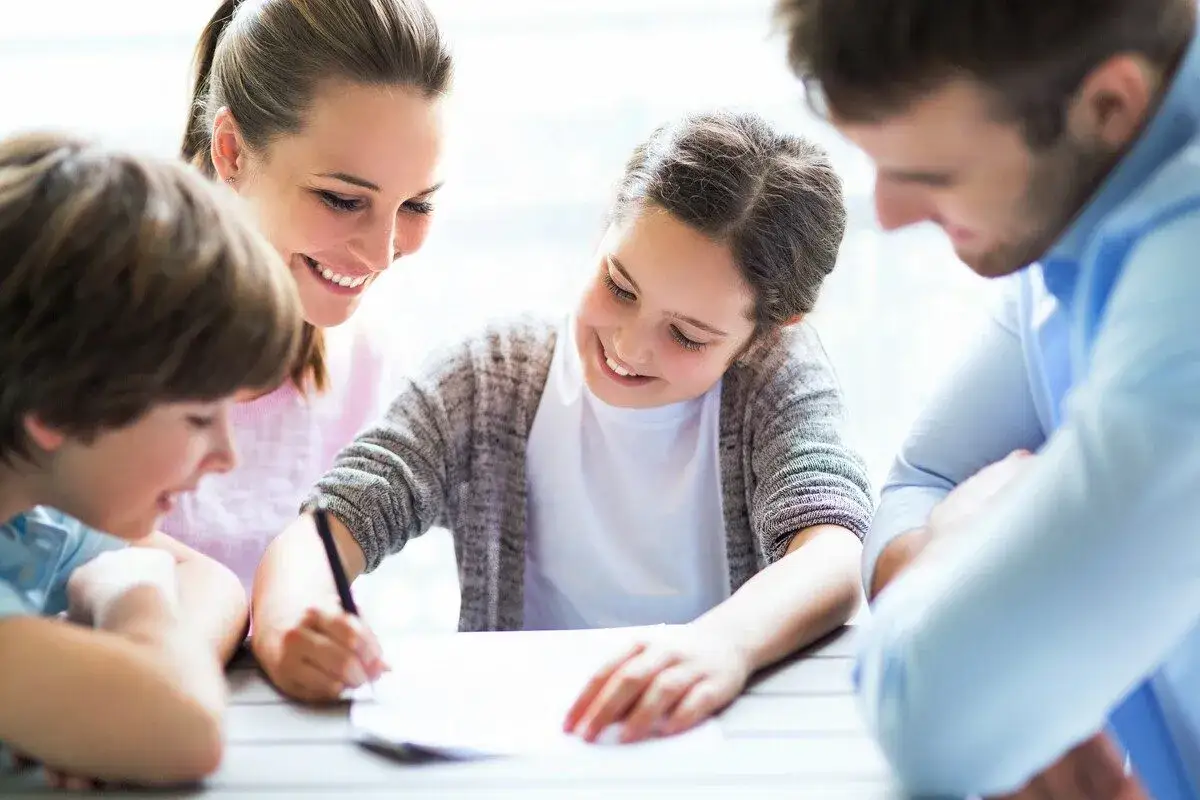 Rodzina wspólnie odrabia lekcje. Mama z uśmiechem patrzy na córkę piszącą coś na kartce, a tata i syn przyglądają się z zainteresowaniem. To miłe zdrobnienie rodzinnych chwil.