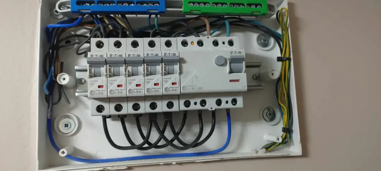 Widok na starą instalację elektryczną w białej obudowie. Widać bezpieczniki, przewody i złącza.