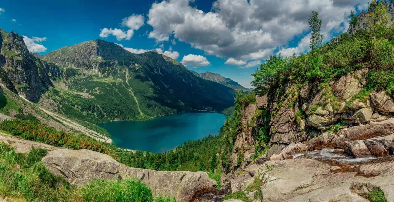 Widok na turkusowe jezioro otoczone zielonymi zboczami i skalistymi szczytami Tatr Wysokich.