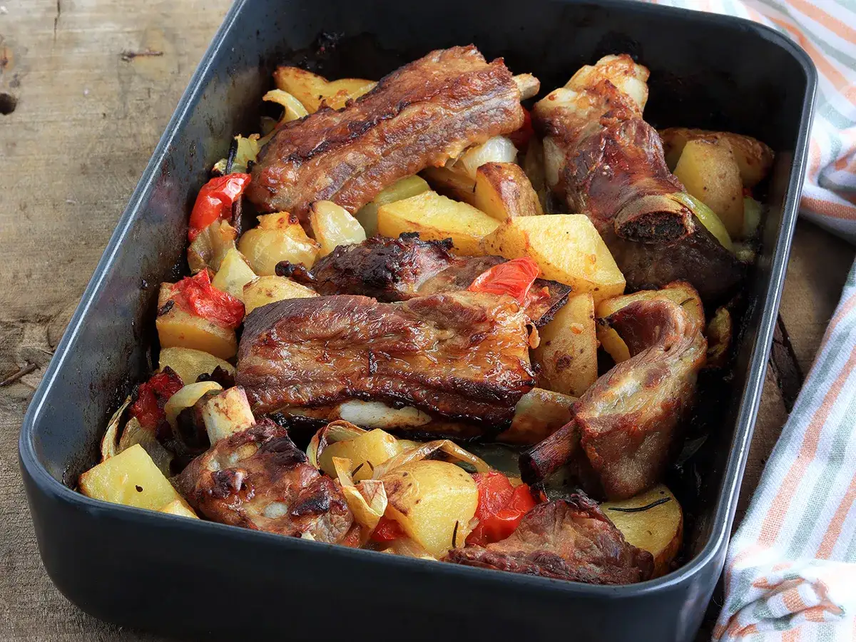 Succulente costine al forno con patate, peperoni e cipolle in una teglia scura, pronte per essere gustate.