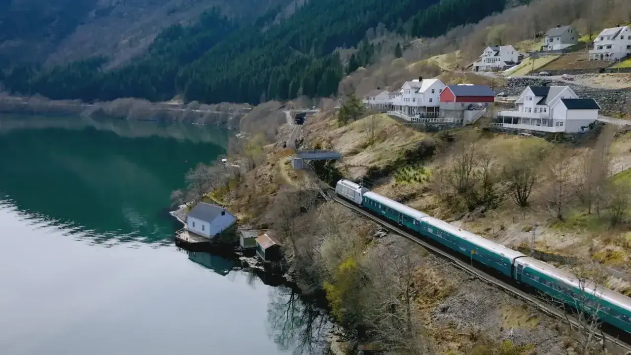 Ein Zug fährt entlang eines Fjords, auf dem Weg von Bergen nach Oslo. Häuser schmiegen sich an den Hang.