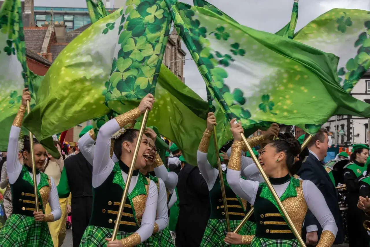 Radosny przemarsz z zielonymi flagami w koniczynki na Święto Świętego Patryka. Dziewczyny w błyszczących strojach tańczą z dumą.