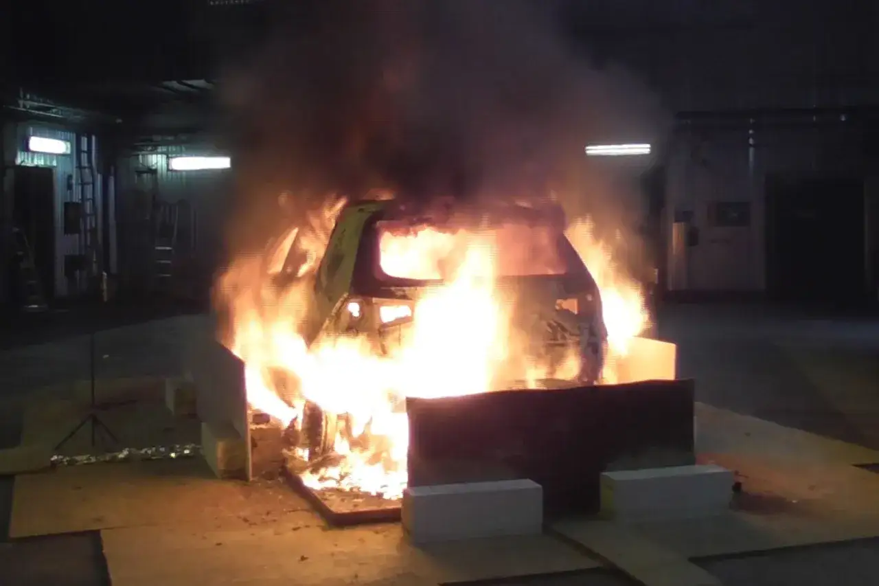 En elbil i ett garage st&aring;r i full brand. L&aring;gorna sl&aring;r upp ur bilen och tjock r&ouml;k fyller rummet. Detta illustrerar brandrisk vid laddning av elbil.