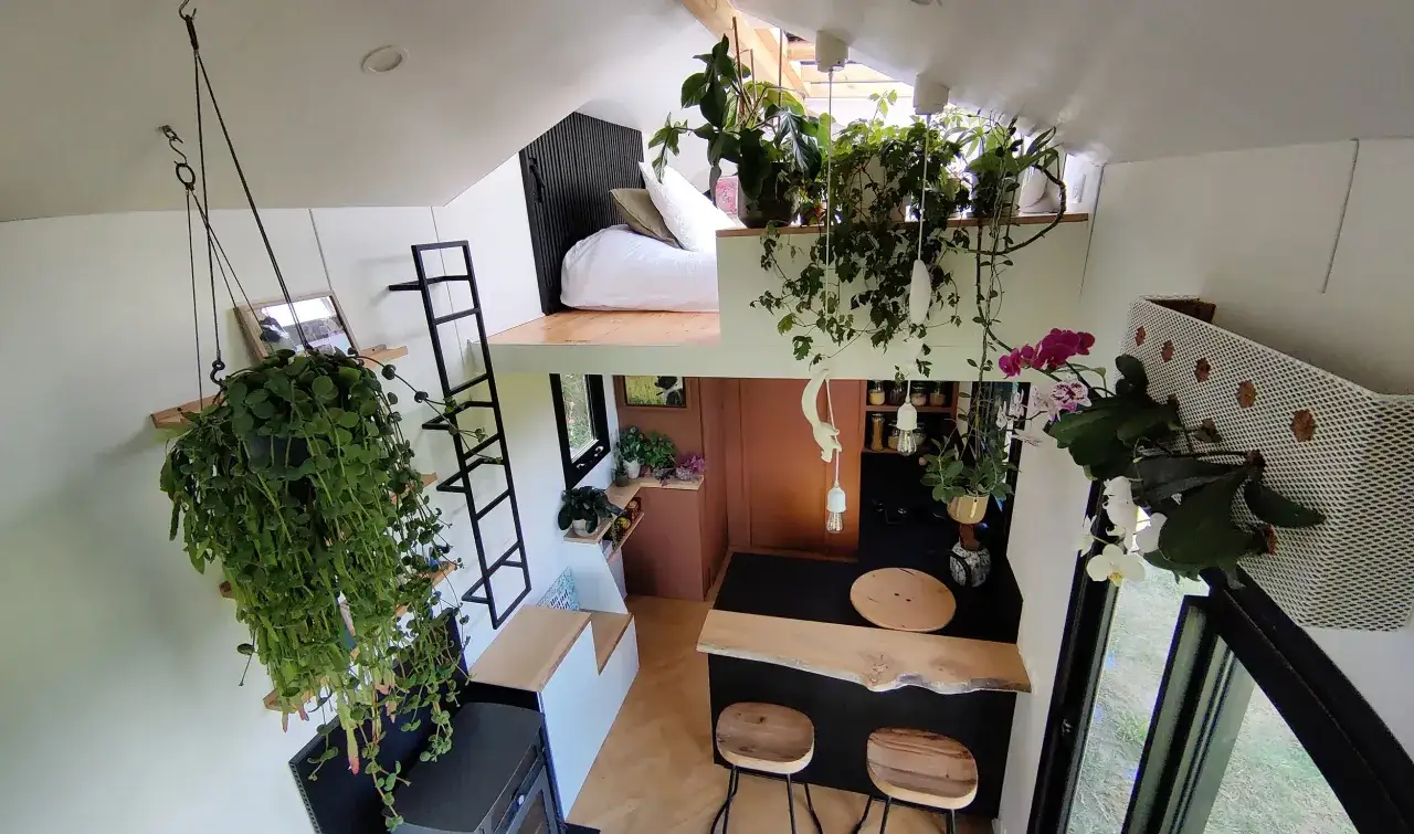 Vue aérienne du **tiny house interieur** luxuriant, avec des plantes suspendues, un lit en mezzanine et un coin cuisine cosy.